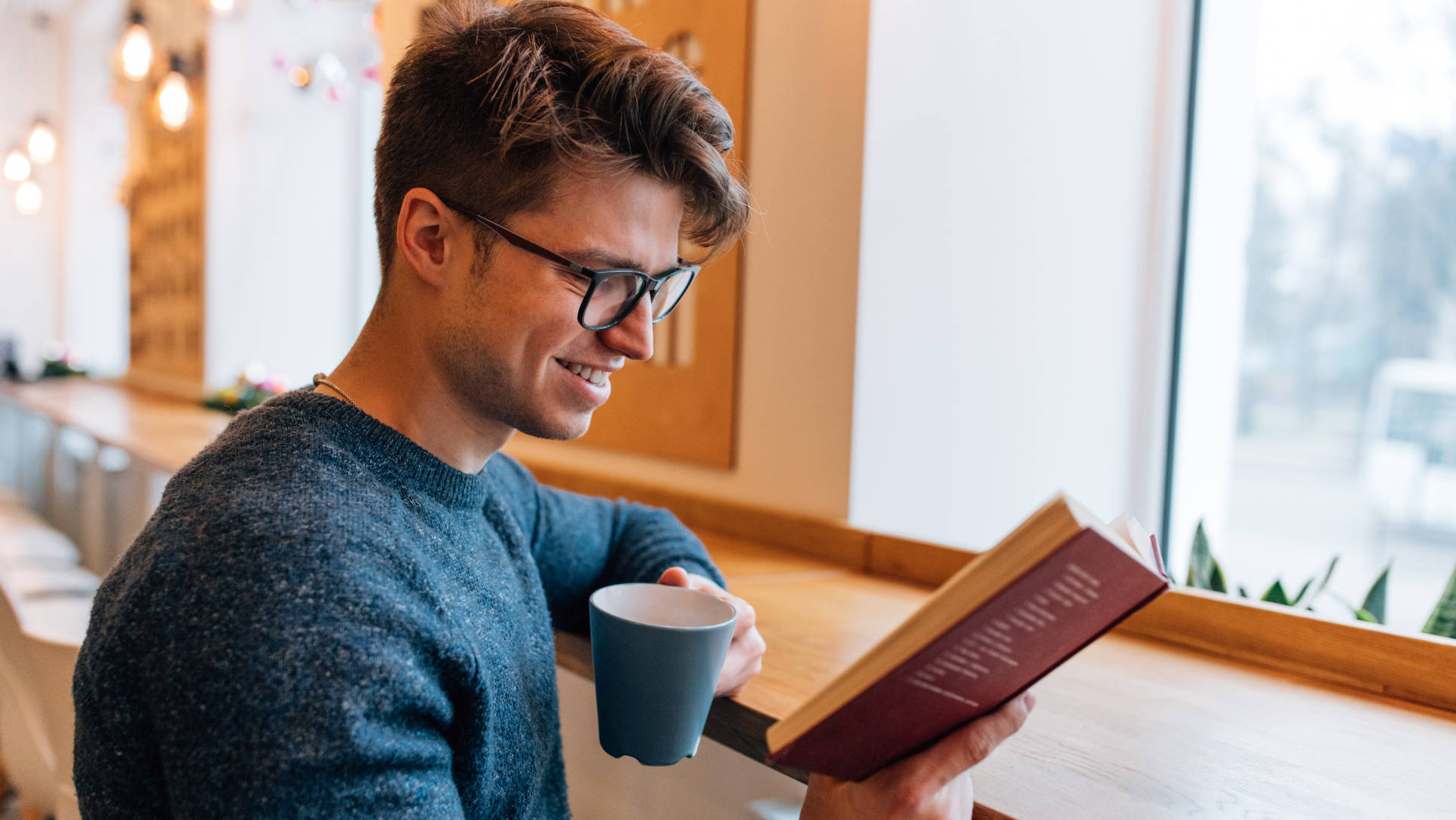 Glad young man resting at cafe, reading interesting book, drinking tea. Dressed in stylish sweater, in eyeglasses.