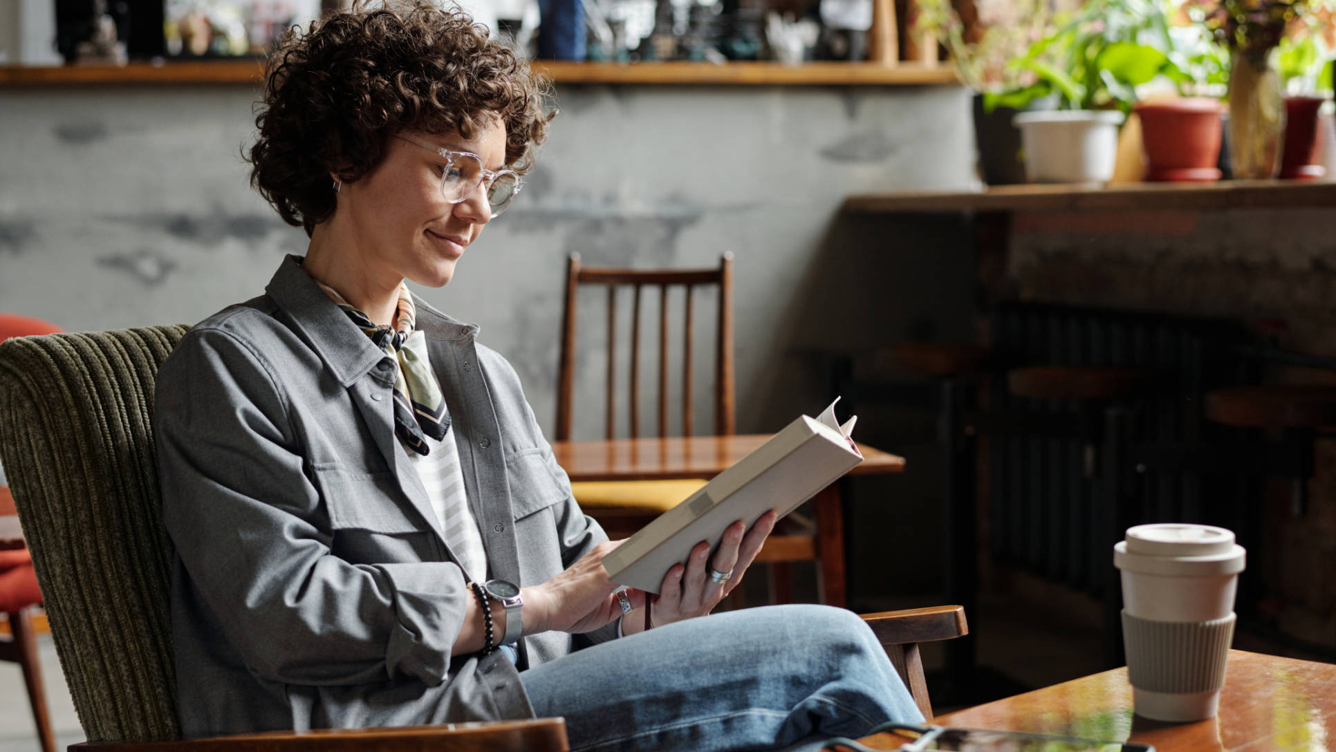 Happy young woman in casualwear and eyeglasses relaxing in armchair with interesting book while spending leisure in cozy cafe